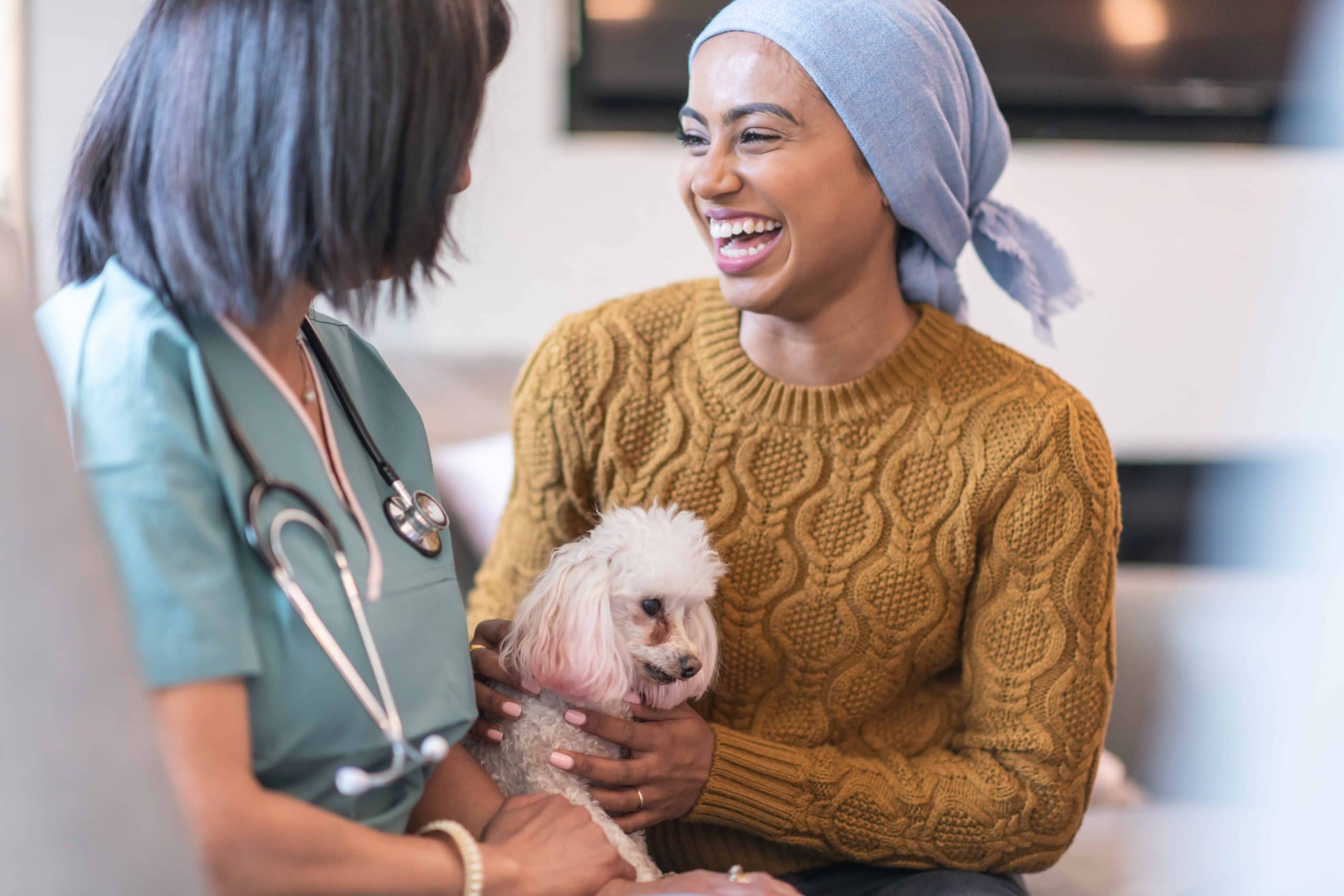 A mixed-race woman with cancer is meeting with her female physician. The patient is laughing while having a conversation with her doctor. She is holding her pet dog on her lap. The patient is wearing a headscarf to hide her hair loss from chemotherapy treatment.